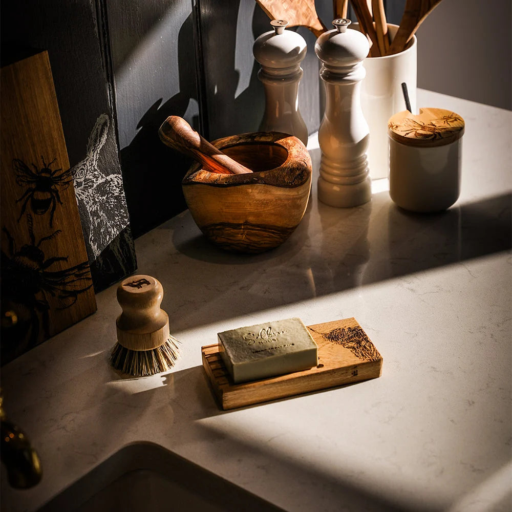 A bar of soap resting on a wooden soap dish on a marble countertop, surrounded by kitchen items like a wooden brush, mortar and pestle, salt and pepper mills, and a wooden spoon holder in warm, dramatic sunlight.