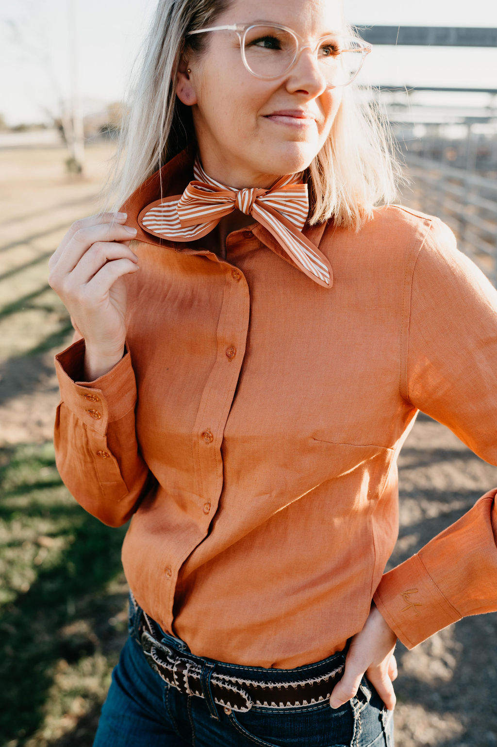 Kitty Fordè Camilla Plain Linen L/S Shirt. Woman in an bronze shirt and blue jeans sitting in a field. Woman in an orange shirt and blue jeans sitting in a field.