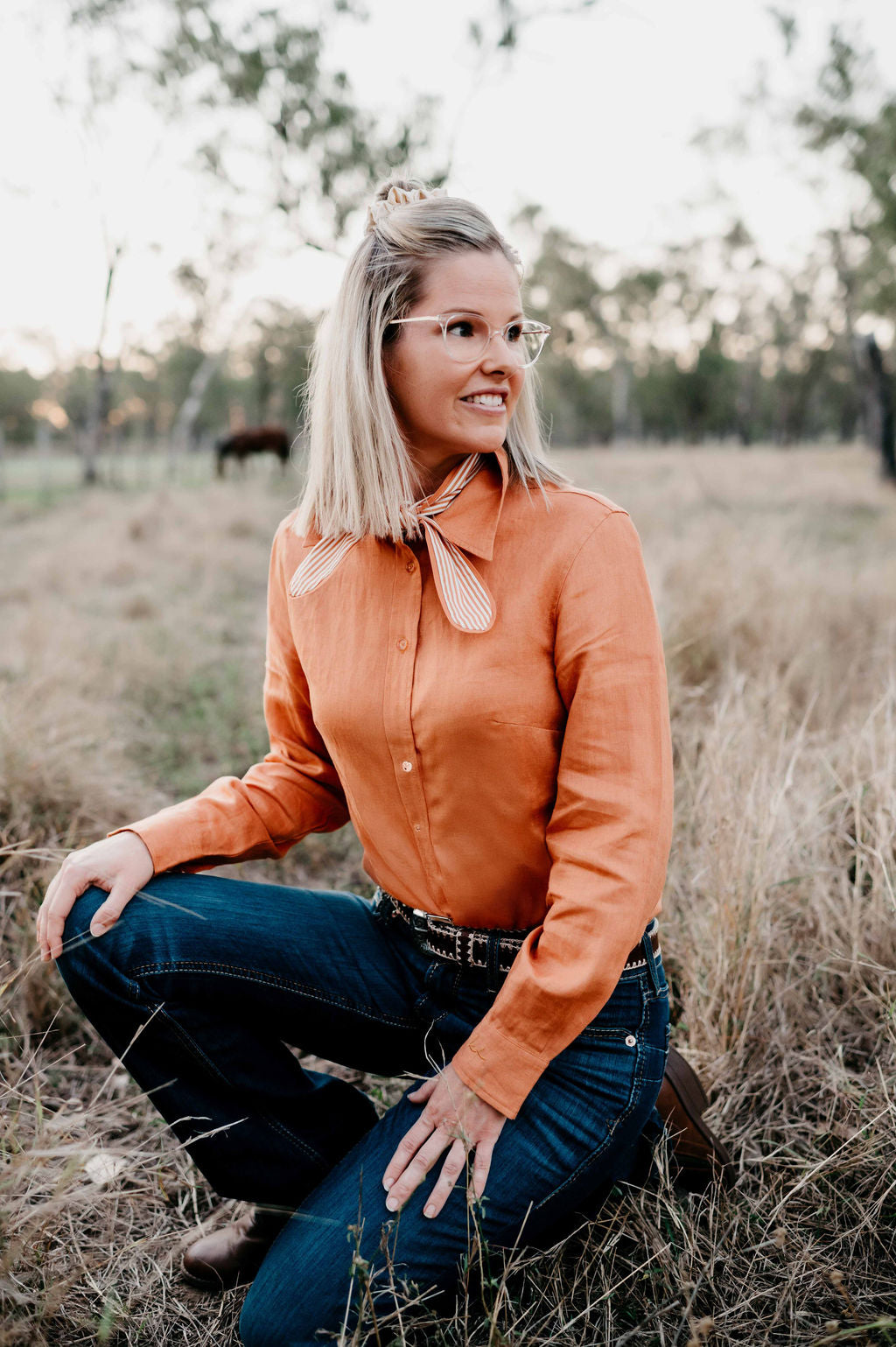 Kitty Fordè Camilla Plain Linen L/S Shirt. Woman in an bronze shirt and blue jeans sitting in a field. Woman in an orange shirt and blue jeans sitting in a field.