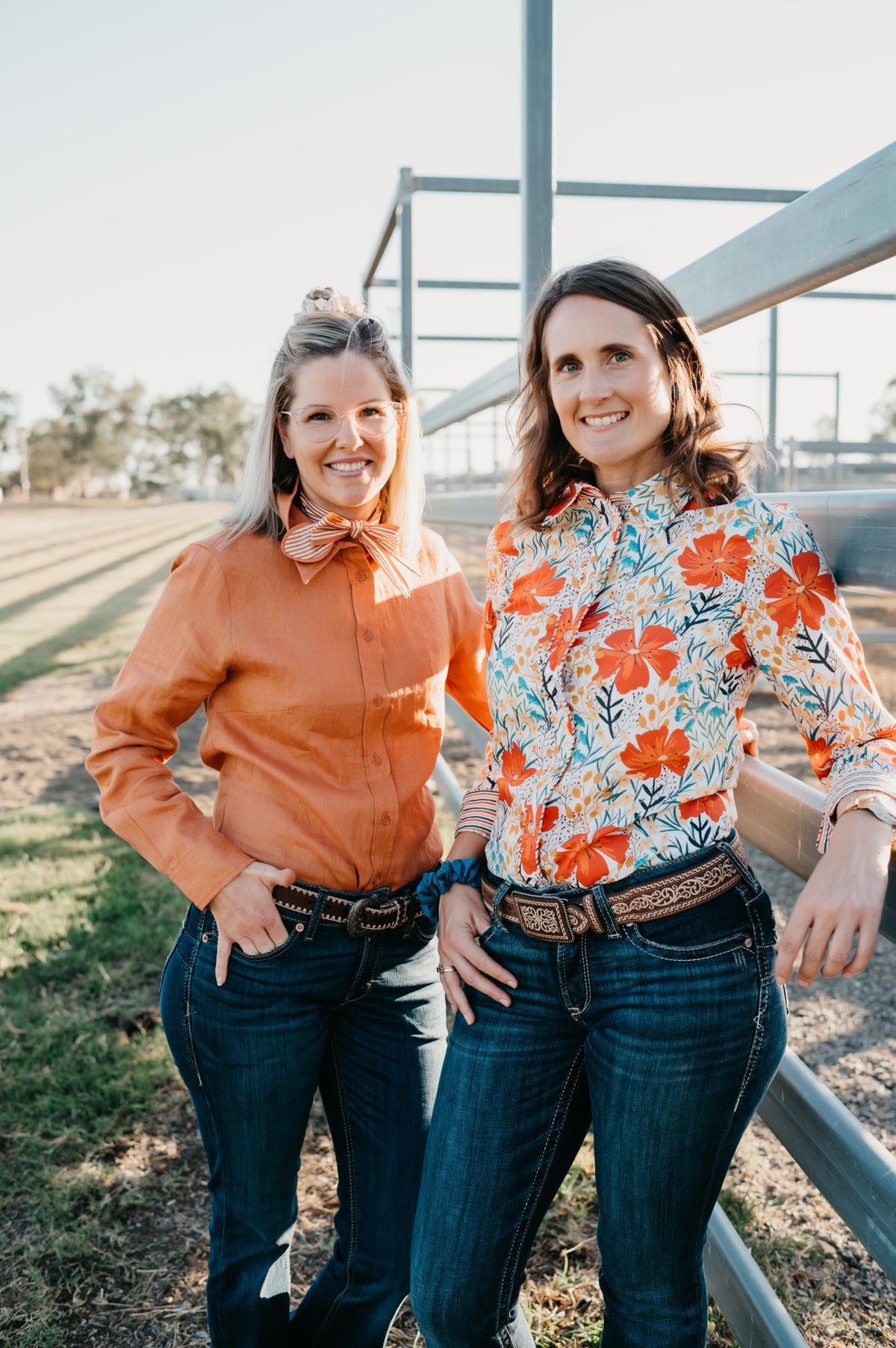 Two women standing outdoors near a metal fence, wearing colorful shirts and jeans.