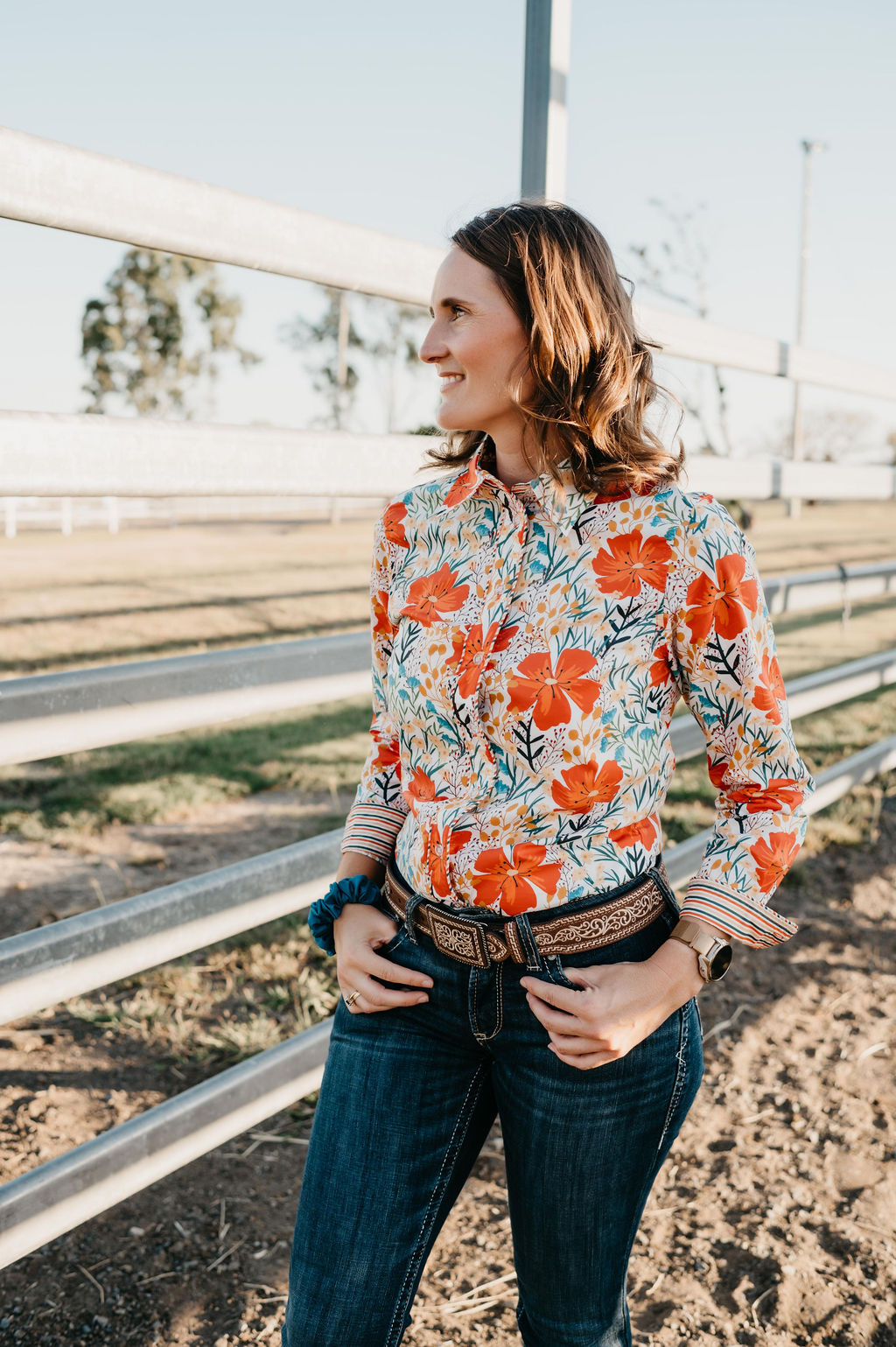 Kitty Fordè Margie Printed L/S Shirt. Woman wearing a colorful floral shirt and jeans standing near a metal fence.