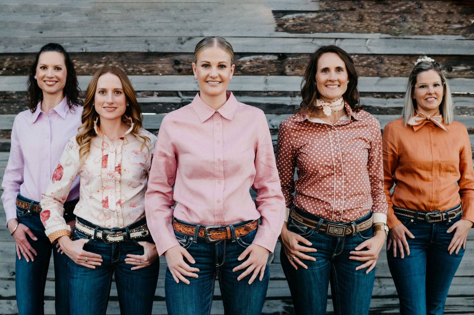 Five women standing together wearing patterned shirts and jeans against a wooden background