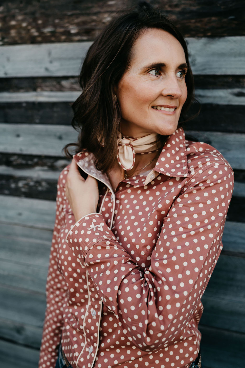 Kitty Fordè Petrina Printed L/S Shirt. Woman wearing a brown polka dot blouse and jeans against a wooden wall.