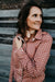 Kitty Fordè Petrina Printed L/S Shirt. Woman wearing a brown polka dot blouse and jeans against a wooden wall.