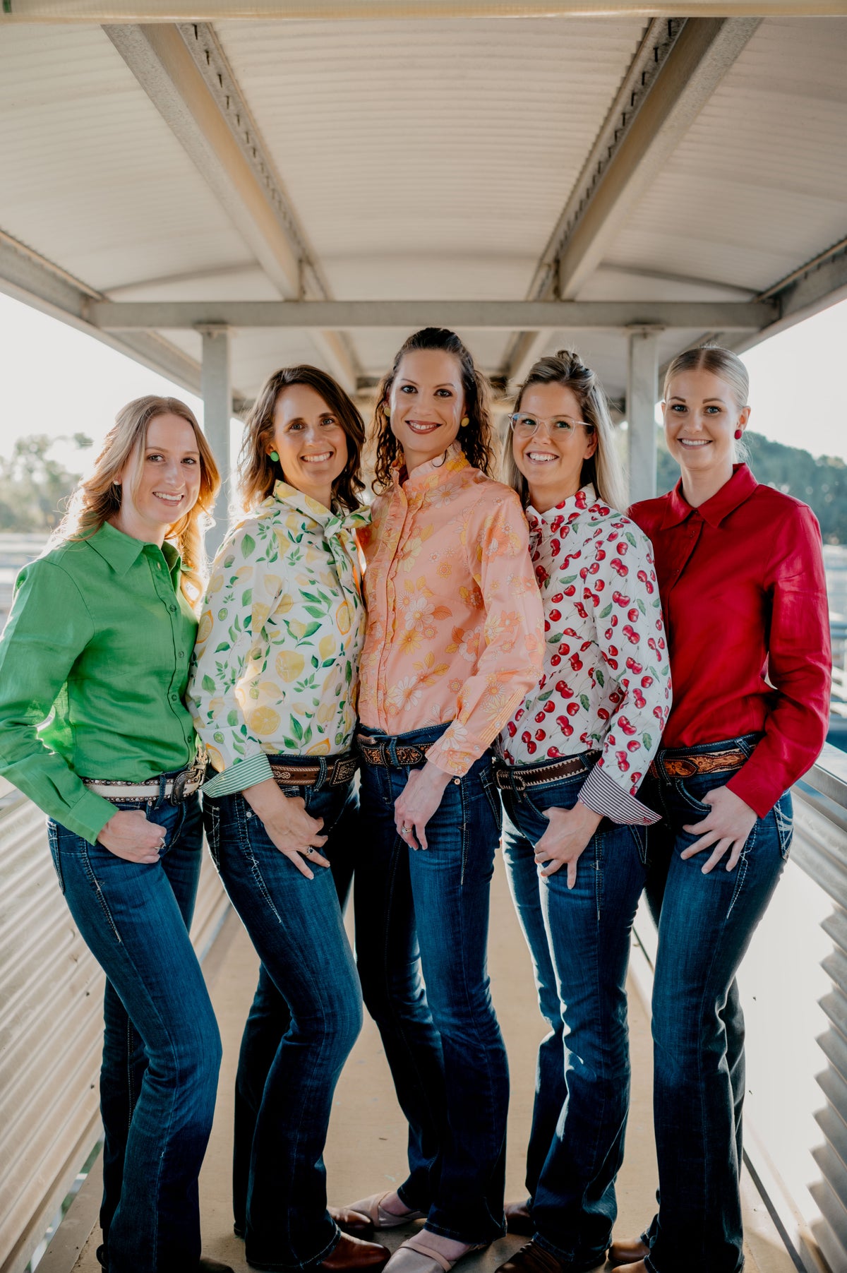 Five women standing together on a wooden dock with a scenic background.