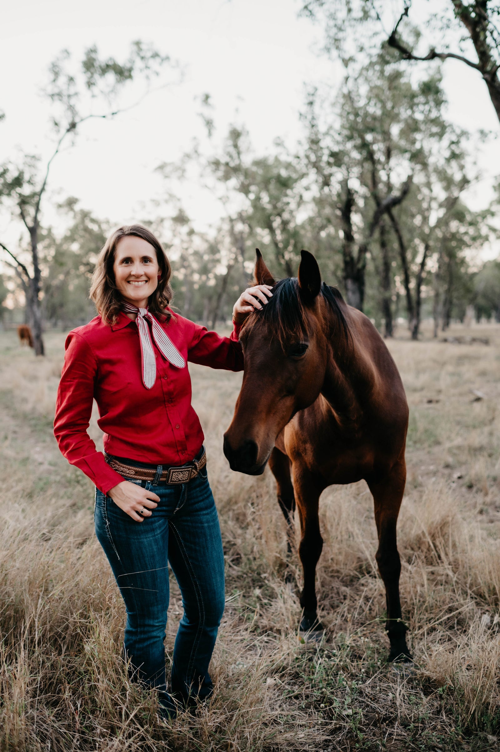 Woman in a red shirt standing next to a brown horse in a field with trees in the background