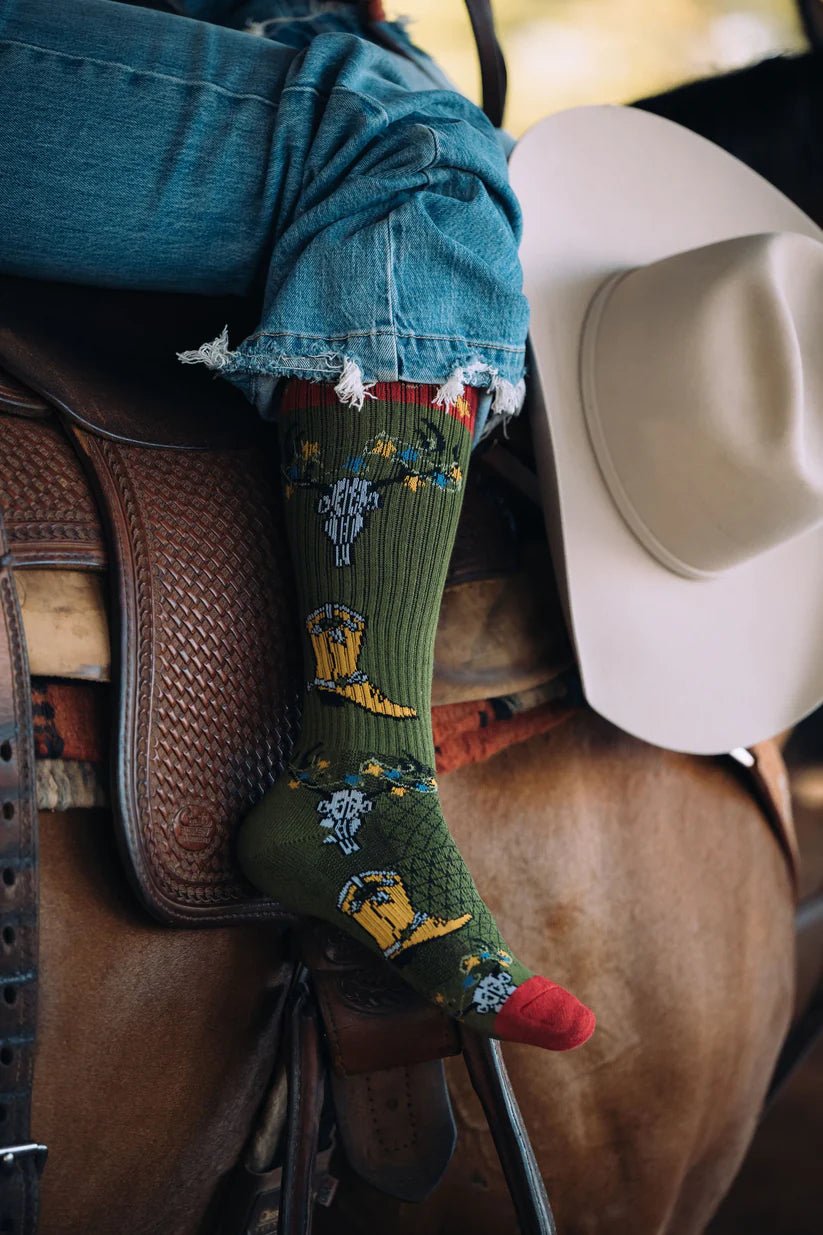 Lucky Chuck Get a Little Western Performance Socks. Close-up of a person wearing green socks with a horse saddle and white cowboy hat in the background.