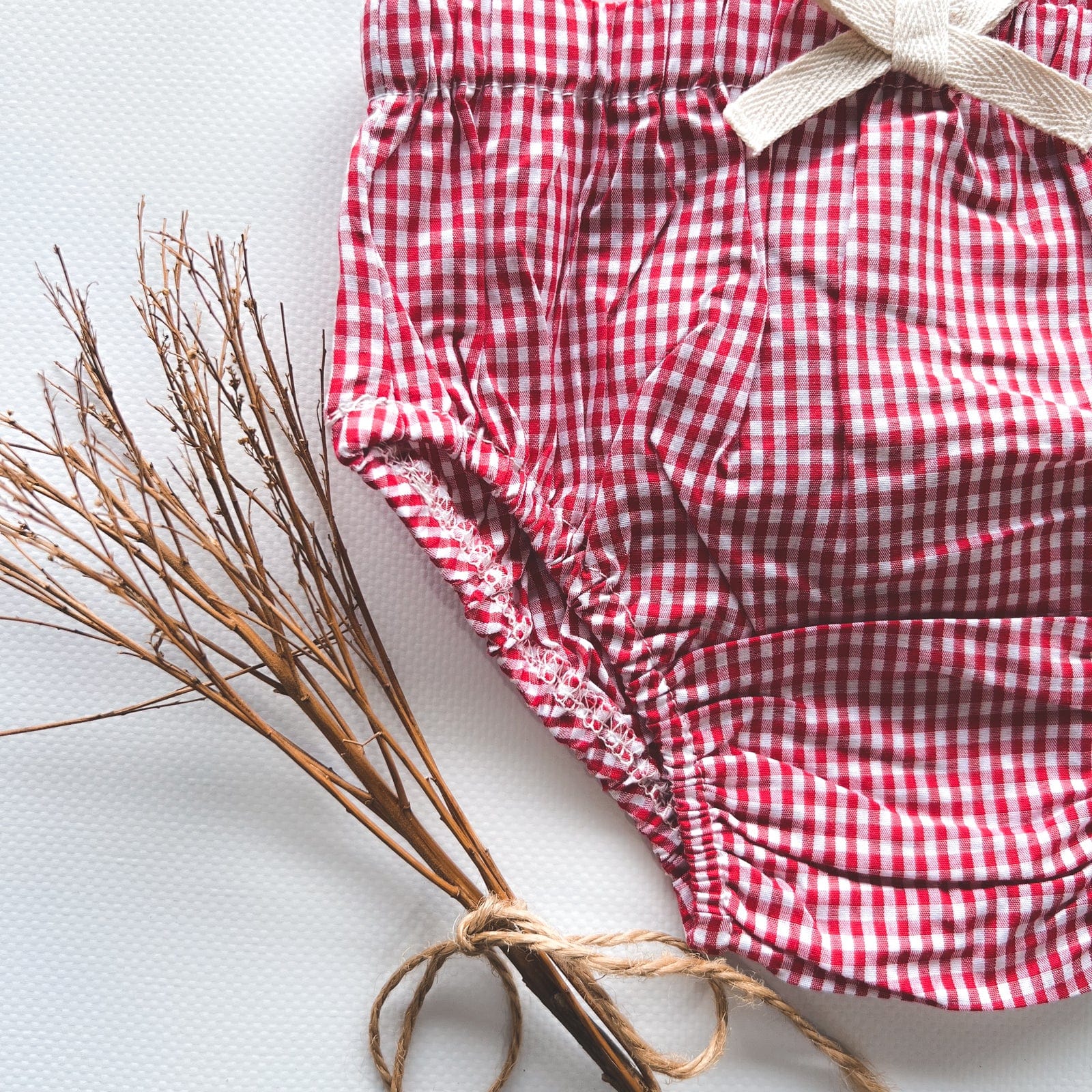 Love Henry Infant Girl's Pilcher. A red and white checkered infant girl's pilcher with a white bow on the front, made of cotton fabric.