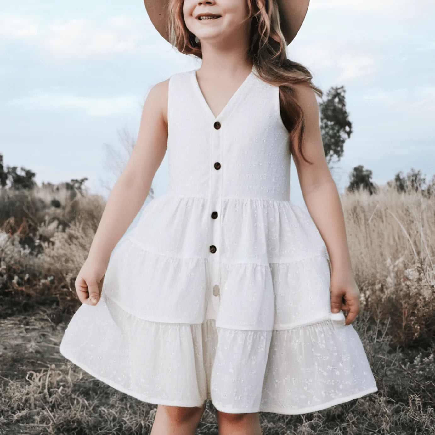 Love Henry Girl's Alice Dress. A young girl wearing a white sleeveless knee-length dress with buttons down the front, standing in a field.