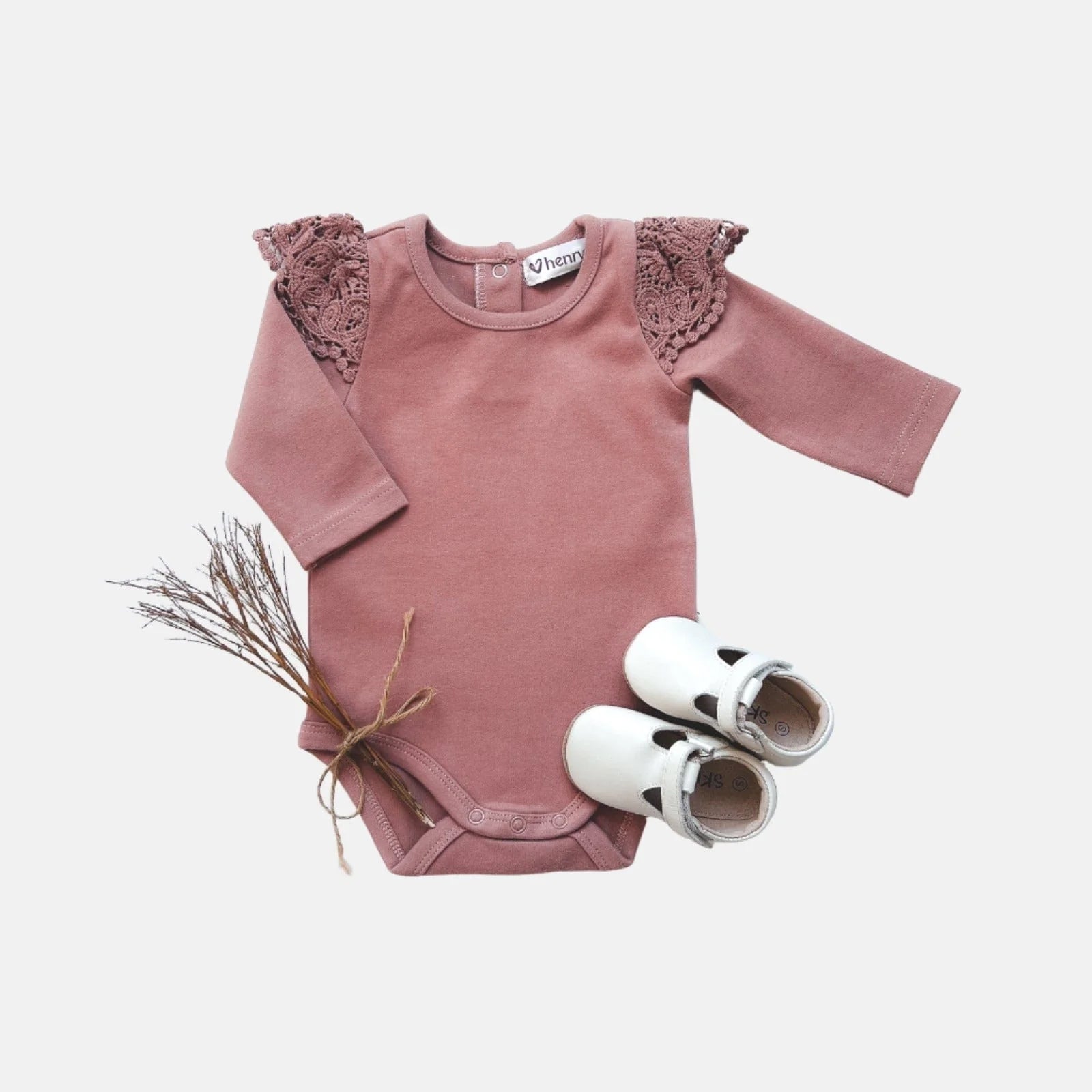 Love Henry Baby Girl's Lace Sleeve Onesie. A pink long-sleeve baby onesie with lace detailing on the shoulders, accompanied by a pair of white baby shoes and a small decorative bundle.