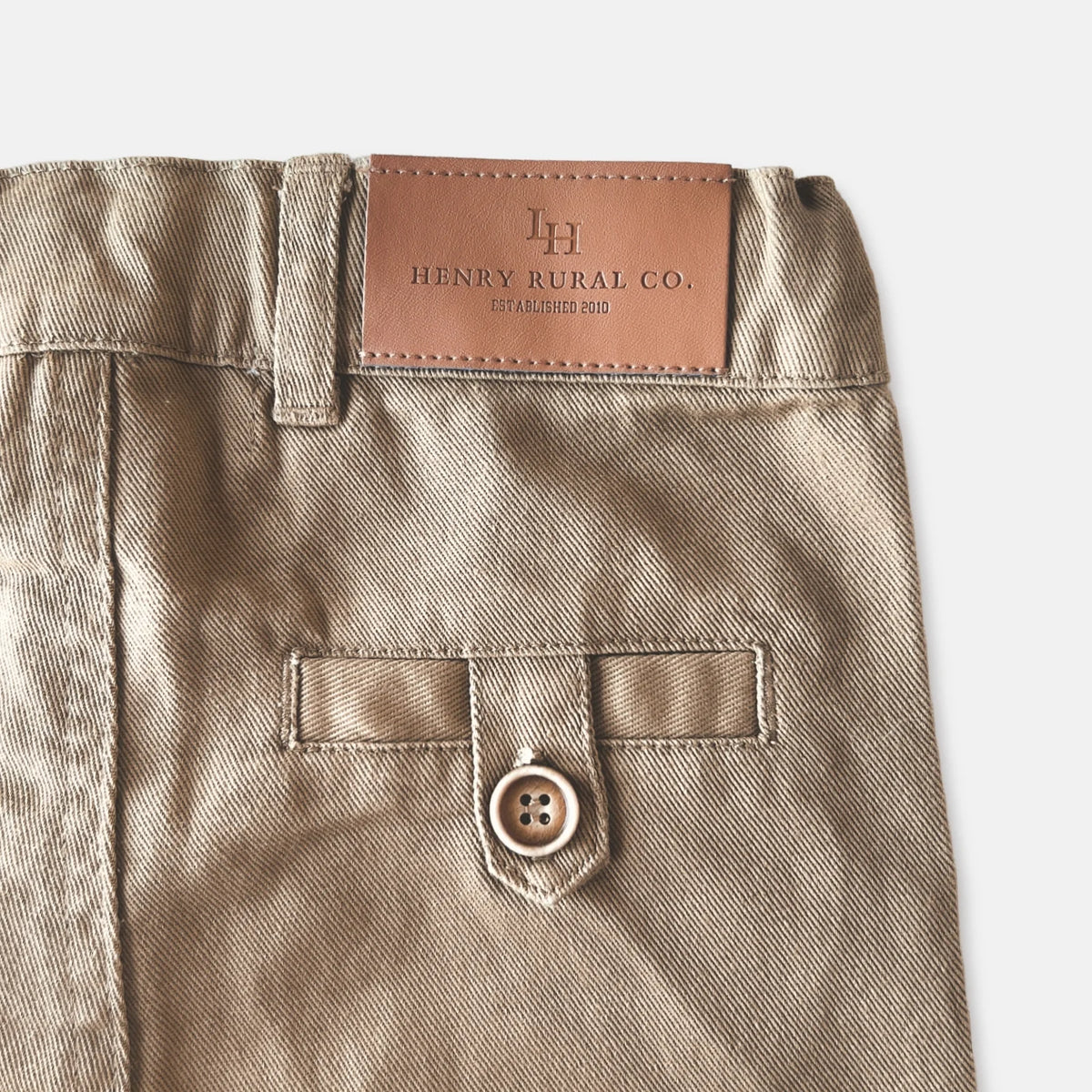 Love Henry Boy's Chino Pants. Close-up of beige pants with a button and brand label on a white background.