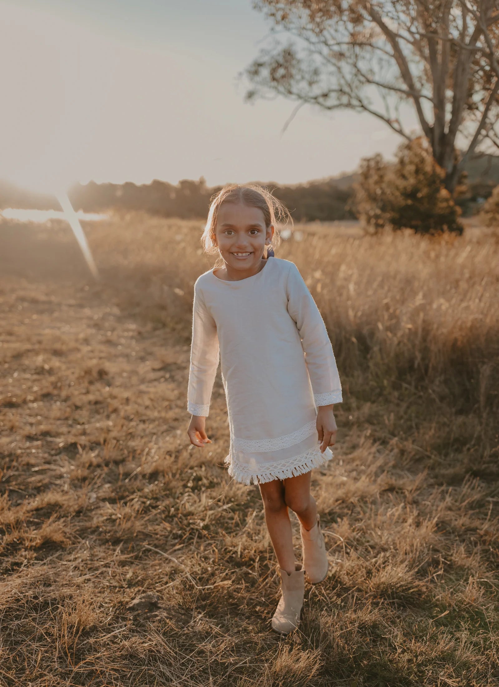 Love Henry Girl's Aleeia Tassle Dress - Linen. Young girl in a white dress standing in a field with a warm, golden light.