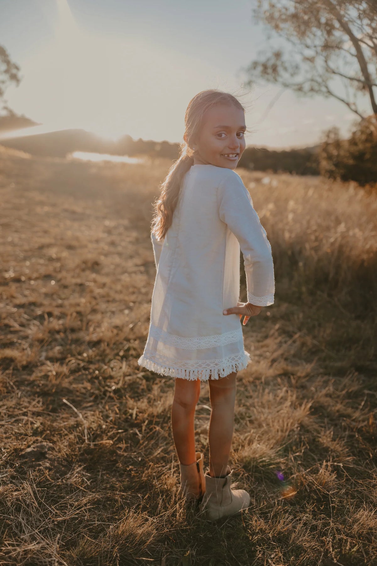Love Henry Girl's Aleeia Tassle Dress - Linen. Young girl in a white dress standing in a field with a sunset in the background