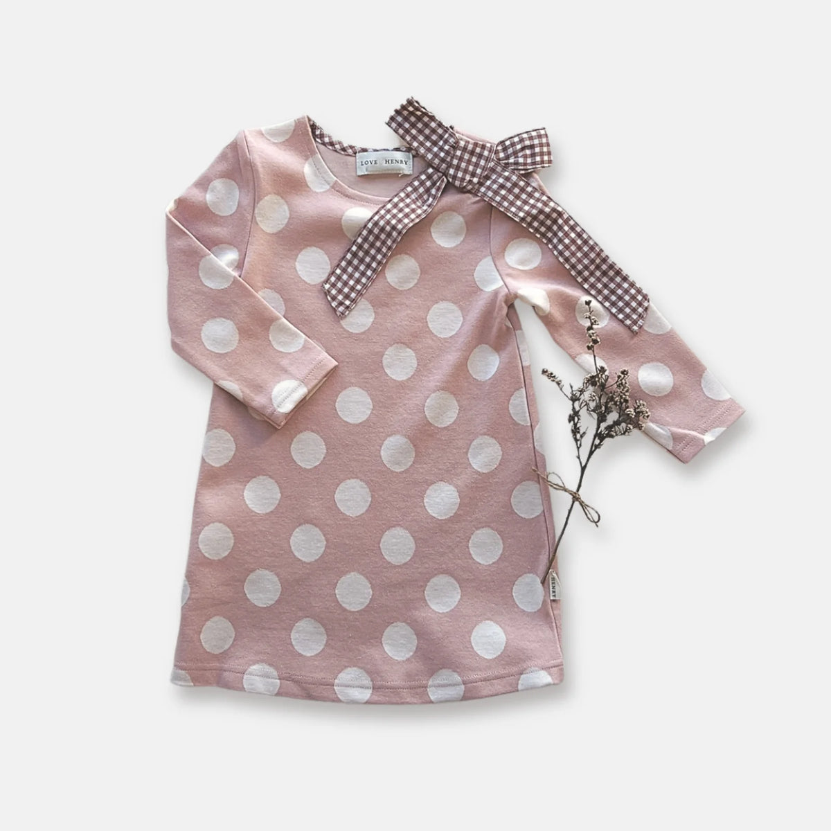 Love Henry Girl&#39;s Mary Dress. A long-sleeved pink dress with white polka dots and a contrasting bow detail at the neckline, presented on a white background with a dried plant to the side.