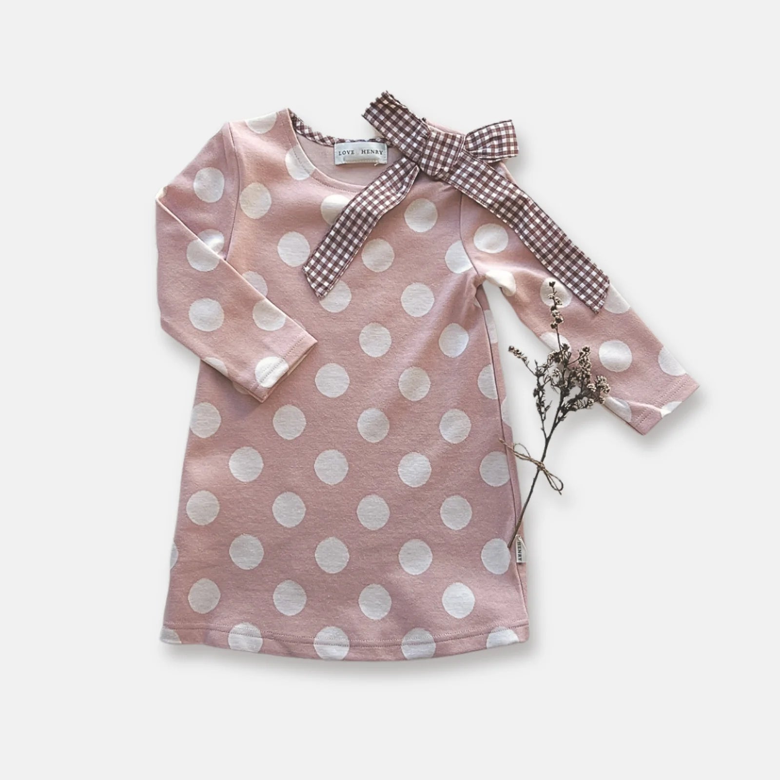 Love Henry Girl's Mary Dress. A long-sleeved pink dress with white polka dots and a contrasting bow detail at the neckline, presented on a white background with a dried plant to the side.