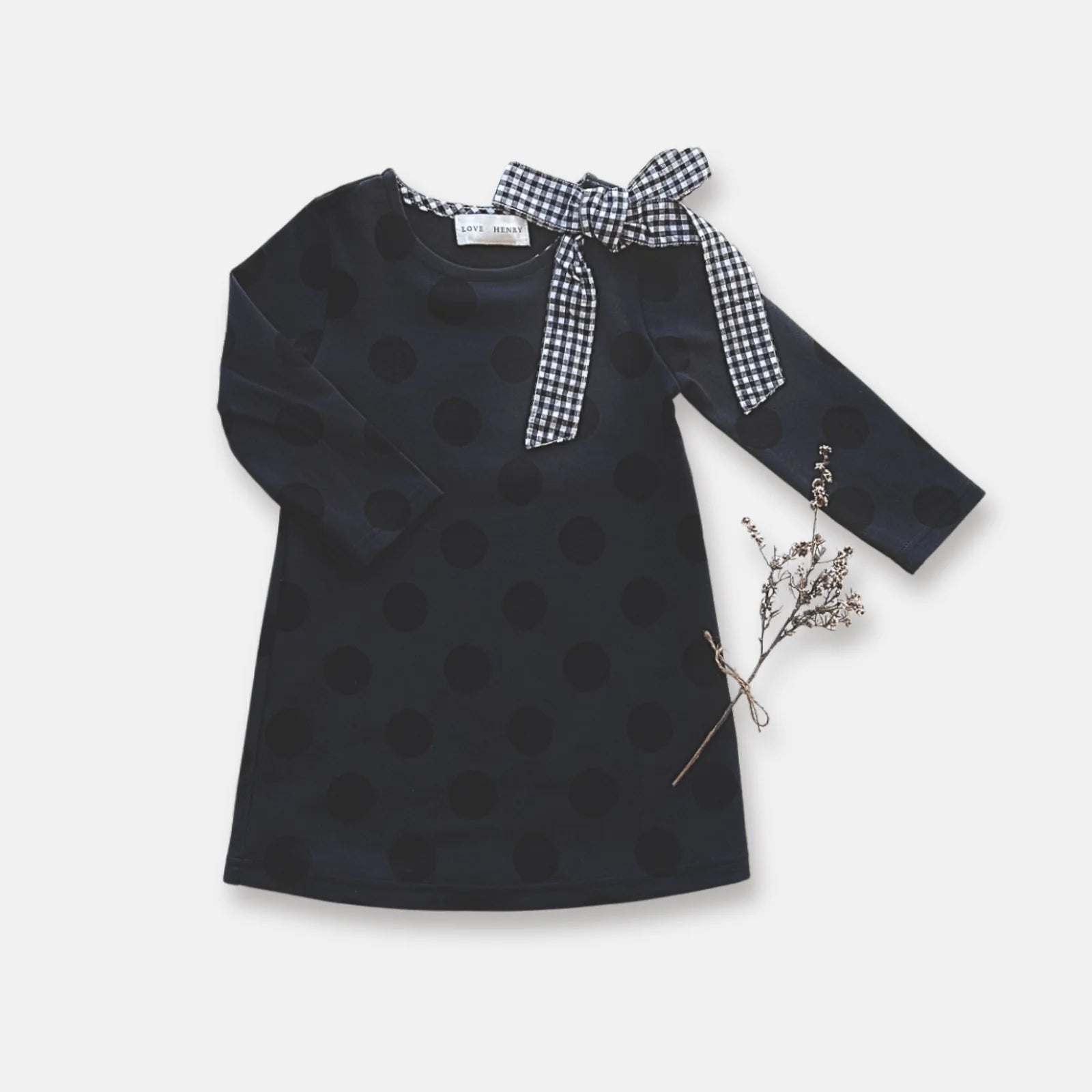 Love Henry Girl's Mary Dress. A navy dress with a polka dot pattern and a contrasting bow detail on the neckline, long sleeves, displayed on a white background with a branch of dried flowers to the side.