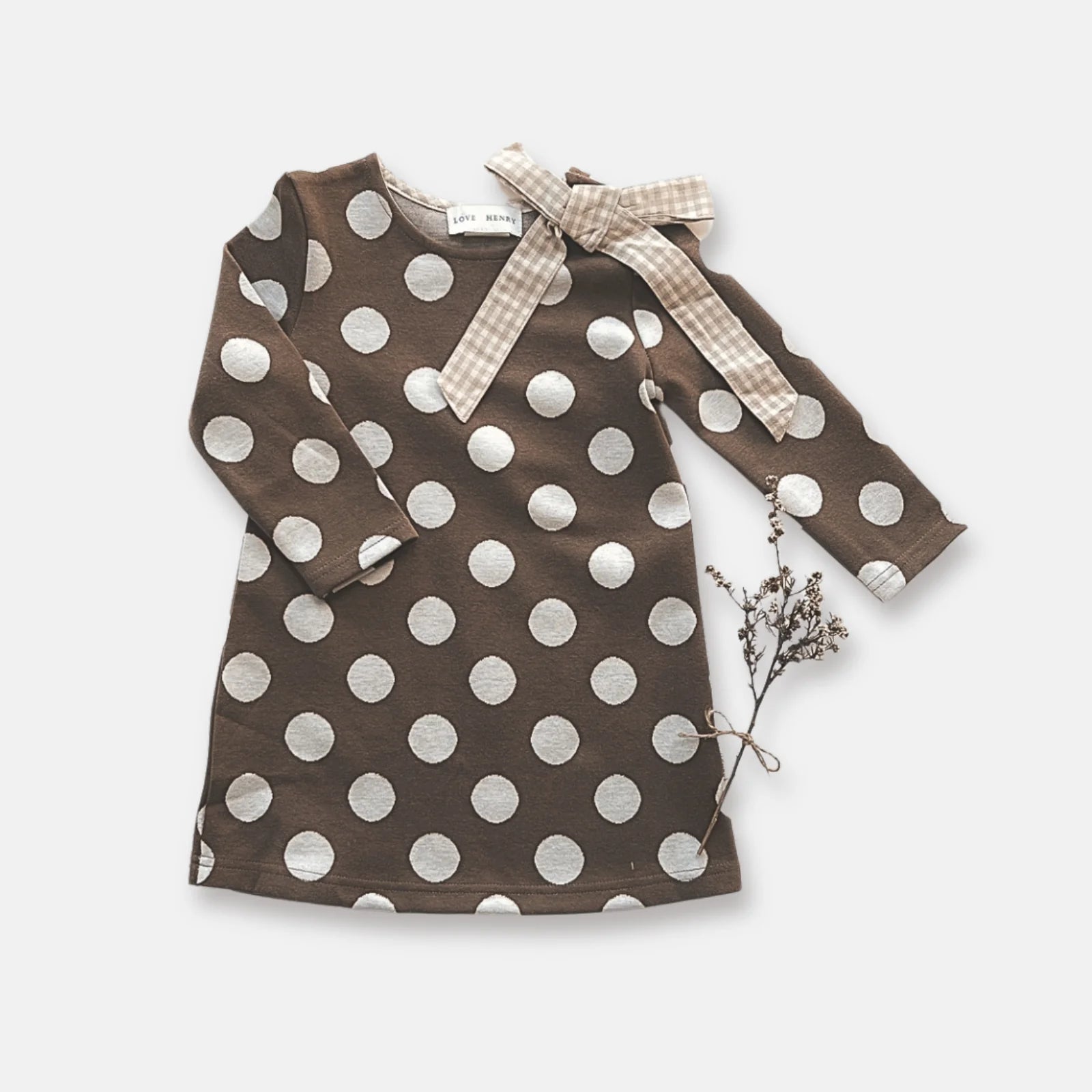 Love Henry Girl's Mary Dress. A long-sleeved brown dress with white polka dots and a contrasting bow detail at the neckline, presented on a white background with a small branch to the side.