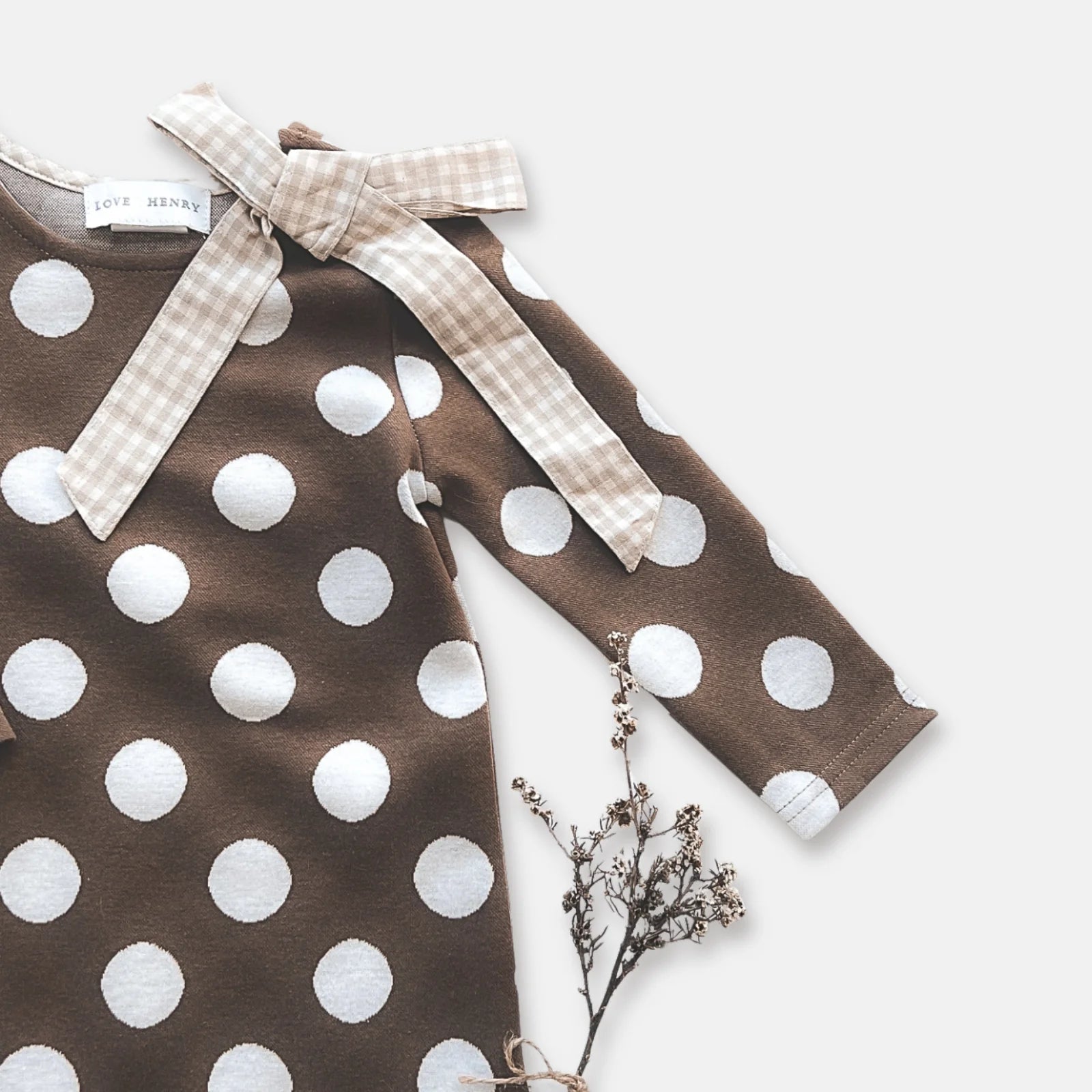 Love Henry Girl's Mary Dress. A long-sleeved brown dress with white polka dots and a contrasting bow detail at the neckline, presented on a white background with a small branch to the side.