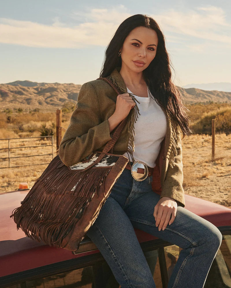 Loyal West Hide And Fringe Large Cowhide Western Handbag. Woman holding a fringed leather bag with a desert landscape in the background