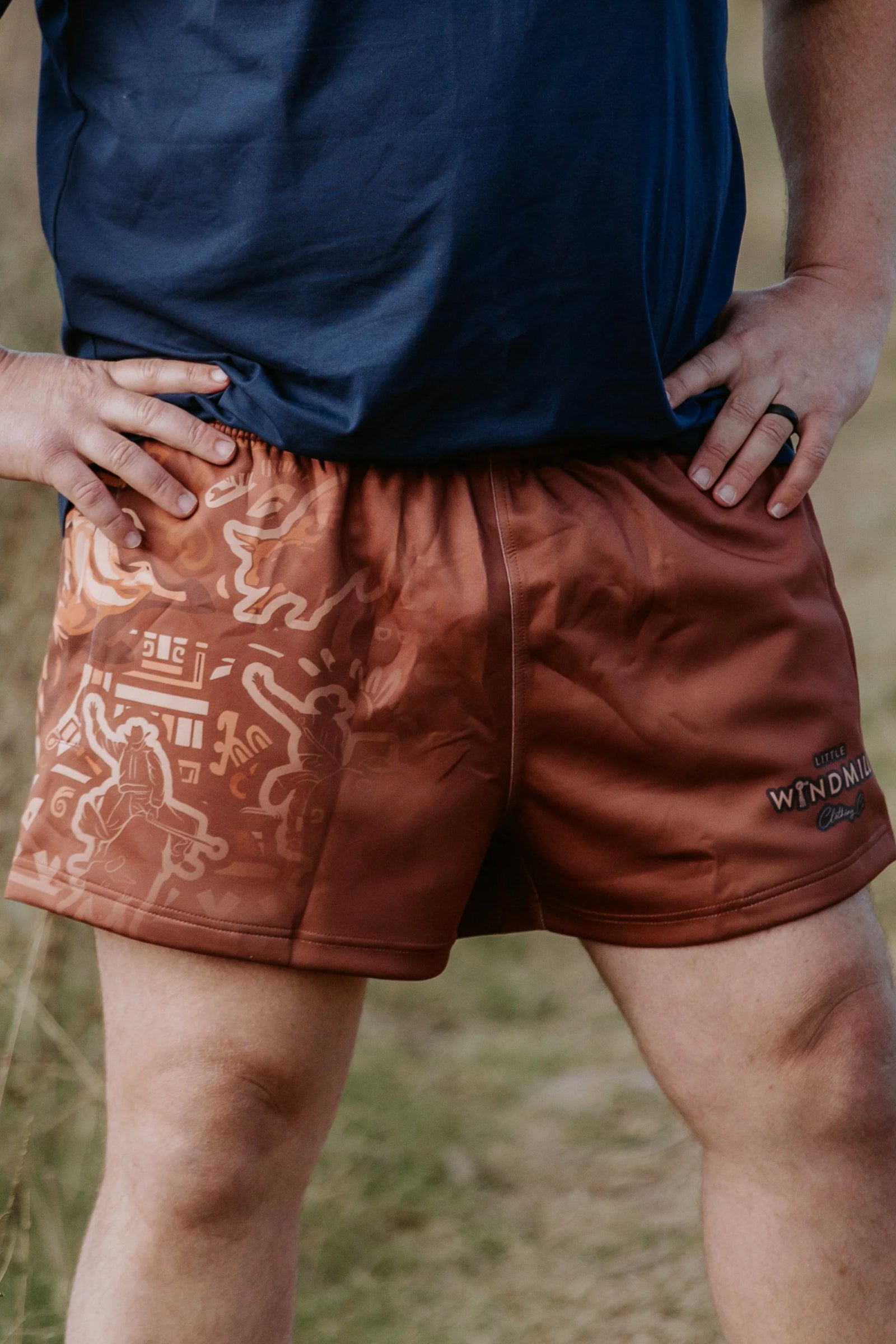 Little Windmill Dust Scene Bull Footy Shorts. Person wearing brown shorts with a pattern and visible brand logo, standing outdoors.
