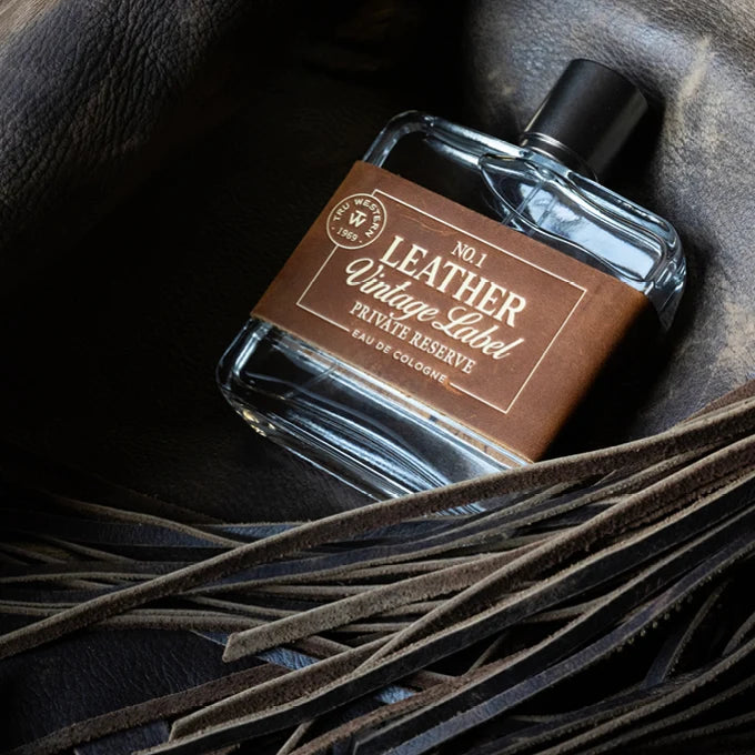 A clear glass perfume bottle with a black cap and a brown label reading 'No.1 Leather Vintage Label Private Reserve Eau de Cologne', styled on a dark leather surface with leather strips in the foreground.