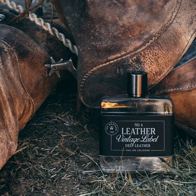 Perfume bottle labeled 'Leather Vintage Label' next to a pair of brown leather boots on a grassy ground.