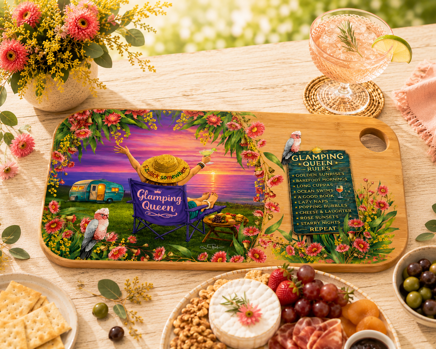 A decorative wooden serving board with a colorful illustrated picnic scene and the words 'Glamping Queen' and a party list, styled on a wooden table with food, flowers, and a drink.