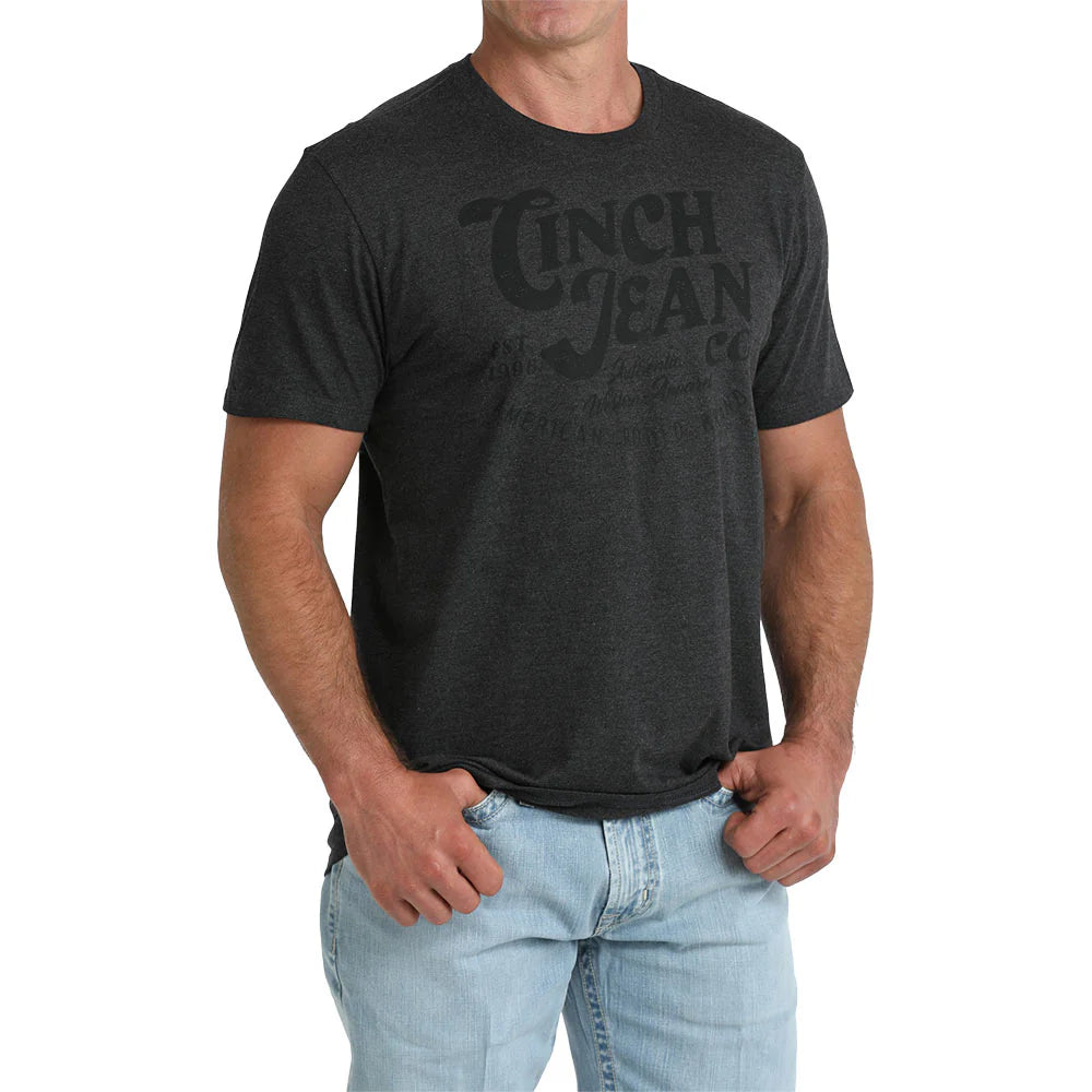 A man wearing a dark charcoal gray heathered t-shirt with a faded black graphic print on the front that reads 'Cinch Jean Co', paired with light blue jeans, against a plain white background.