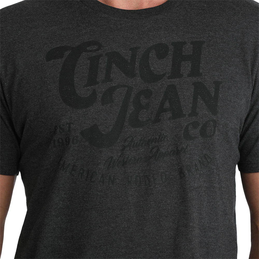 Close-up of a dark charcoal gray heather t-shirt featuring a tonal black distressed graphic print that reads 'CINCH JEAN CO'.