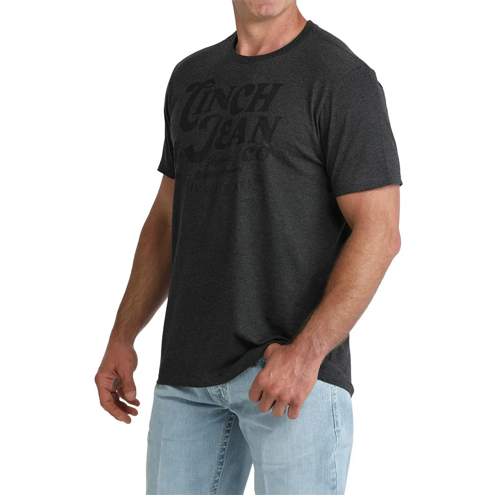 A man wearing a dark charcoal gray heathered t-shirt with a tonal black graphic print of the brand 'Linch Juan'.