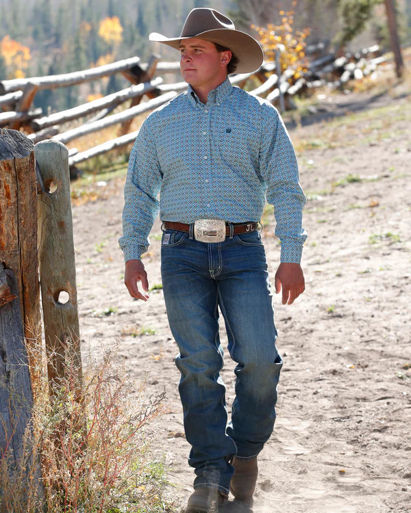 Cinch Men's Classic Fit Geometric Print L/S Western Shirt. Man in cowboy attire walking on a dirt path with wooden fence and trees in the background