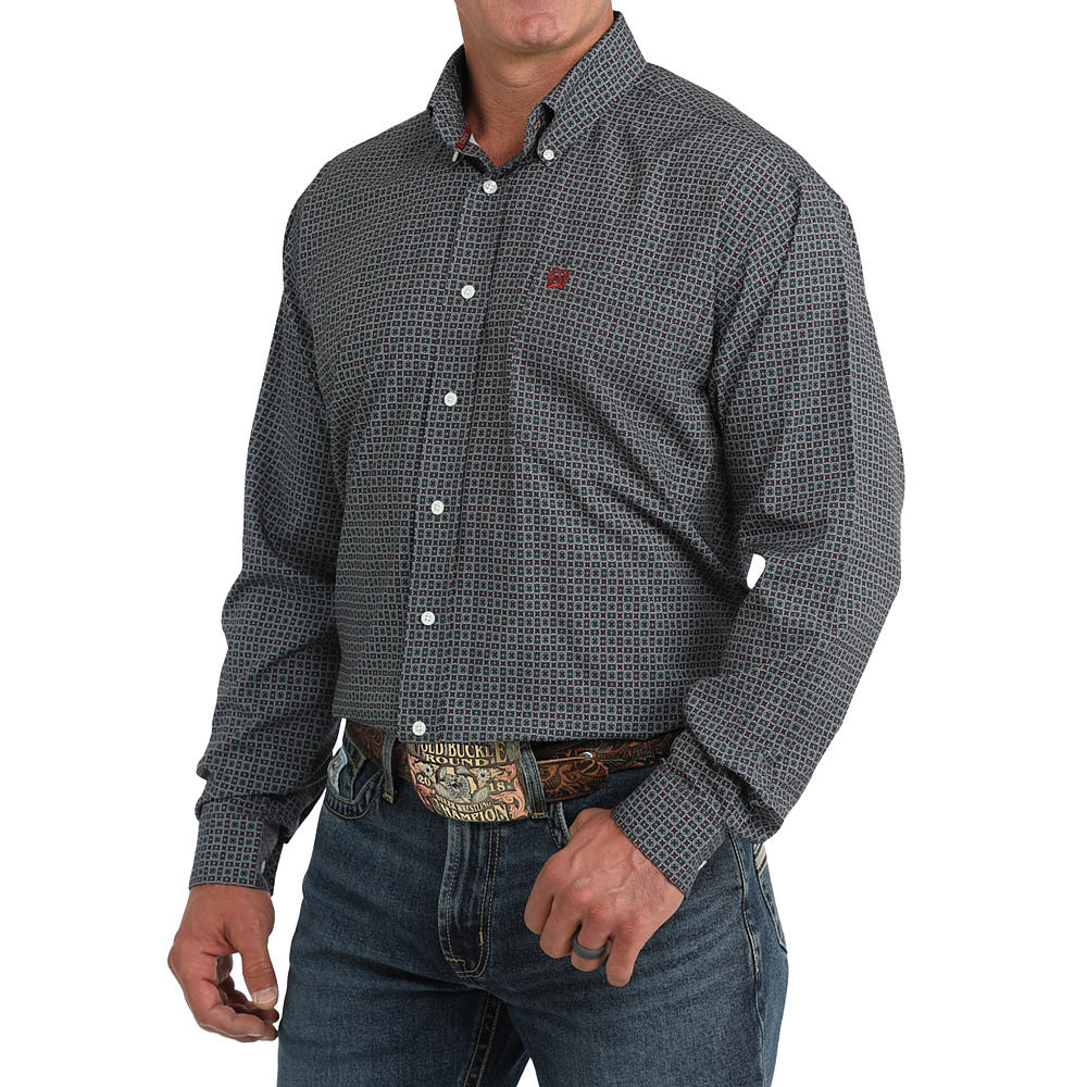 A man wearing a dark gray and black micro-patterned long-sleeve button-down shirt with white buttons, paired with blue jeans and a western belt, against a plain white background.
