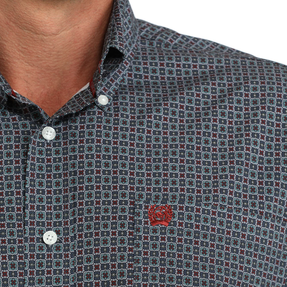Close-up of a man wearing a patterned button-down shirt. The shirt features a small-scale geometric print in navy blue, red, and light blue, with white buttons and a small red embroidered logo on the chest.
