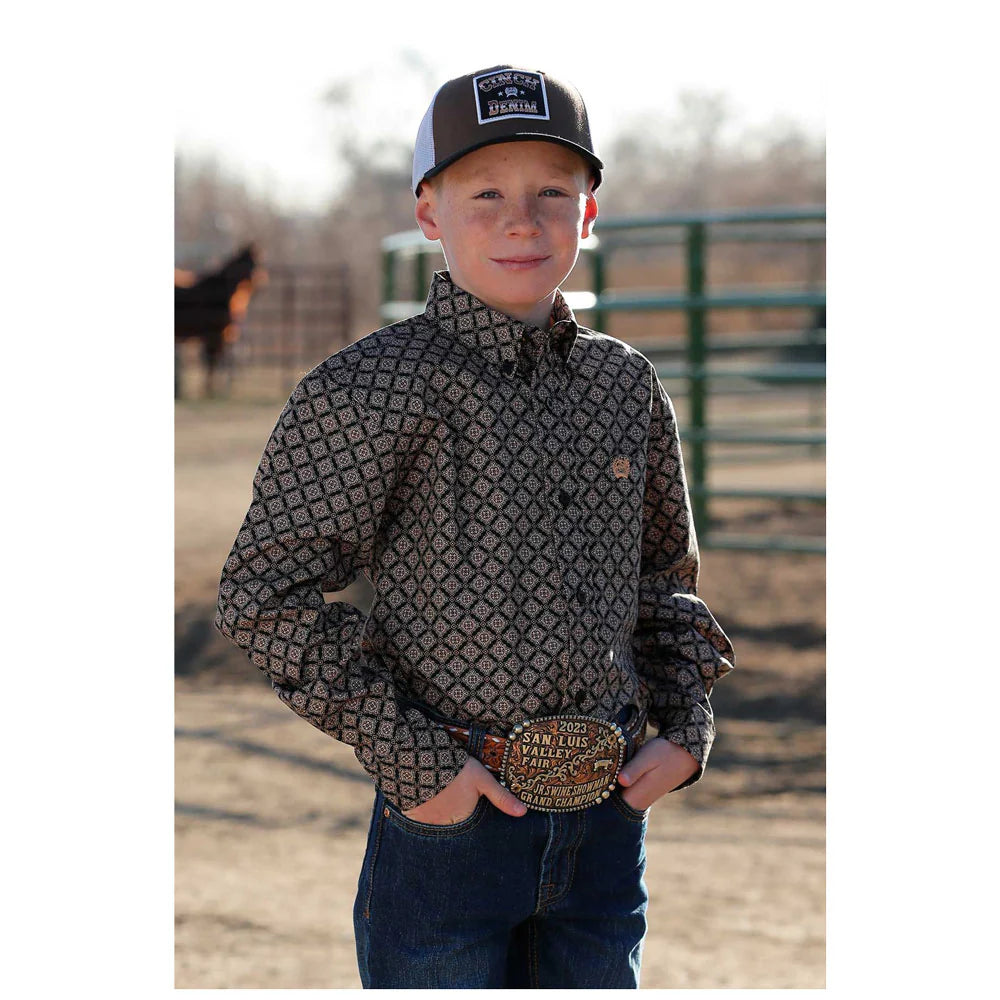Cinch Boy's Medallion L/S Western Shirt. A boy wearing a long sleeve black western shirt with a geometric pattern, button-down collar, and long sleeves. He is also wearing a cap and posing in front of a fence.