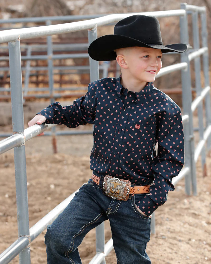 Child wearing a cowboy hat and patterned shirt sitting on a log outdoors.