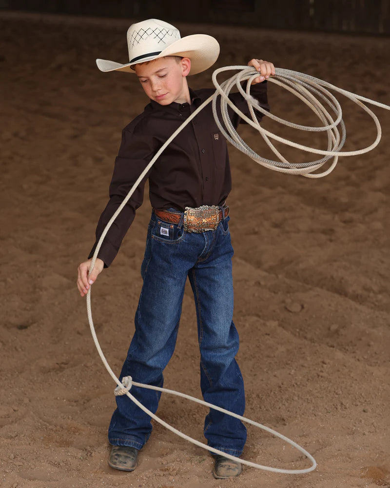 Young boy in cowboy attire with a wide-brimmed hat, brown shirt, and blue jeans.