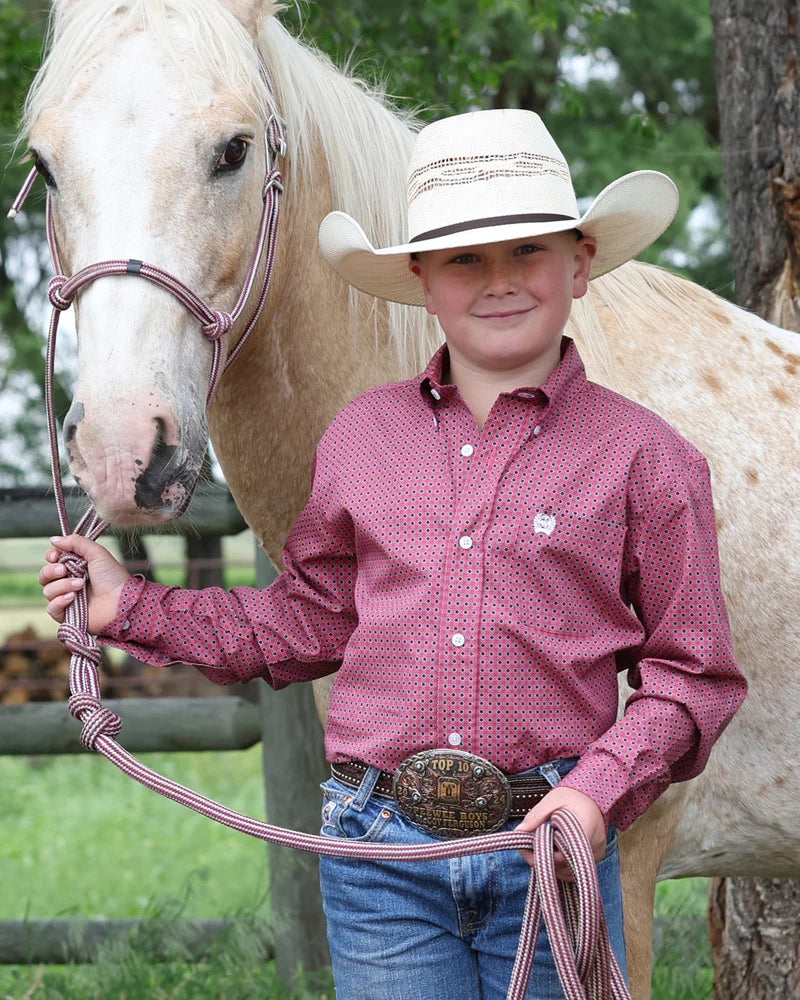 A young boy wearing a western outfit, including a cream cowboy hat, a red patterned button-down shirt, and blue jeans, standing next to a light-colored horse in an outdoor setting.