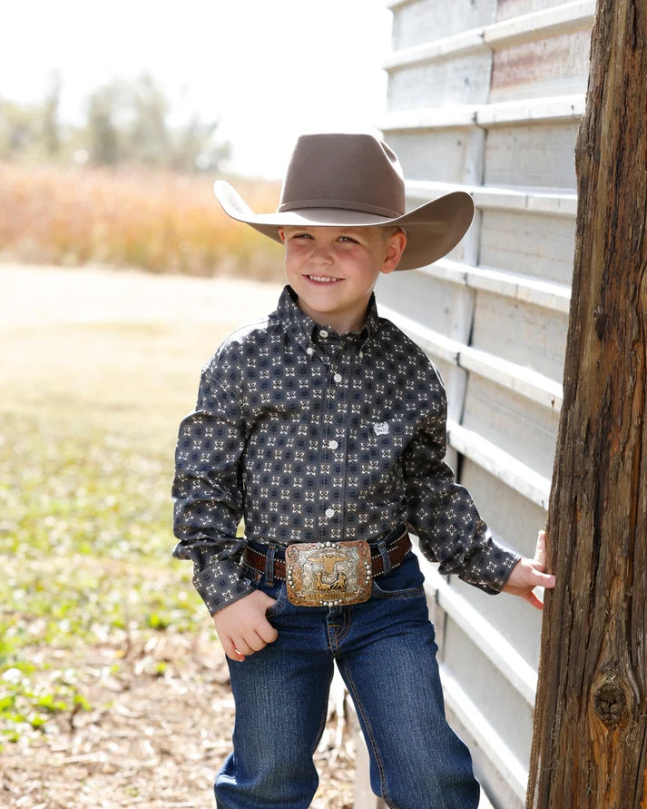 A young boy wearing a brown cowboy hat, a dark patterned long-sleeve western shirt, blue jeans, and a decorative belt stands outdoors next to a corrugated metal wall and wooden post.