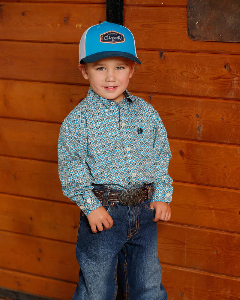 Cinch Boy's Toddler L/S Western Shirt. Child wearing a blue cap and patterned shirt standing against a wooden wall