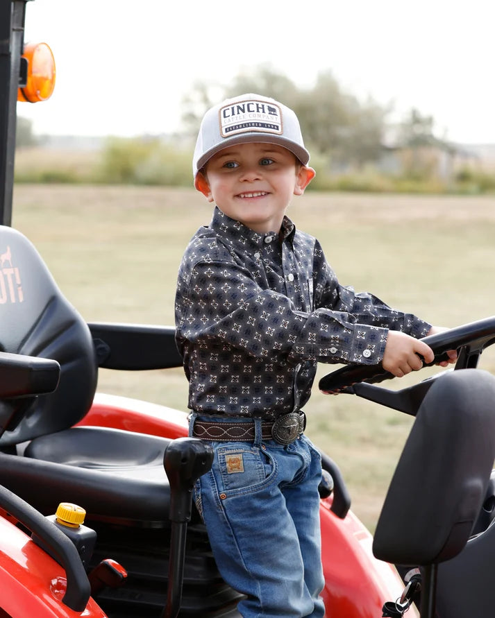 A young boy smiling while sitting in the driver's seat of a red tractor, wearing a black and white patterned long-sleeve shirt, blue jeans, and a white trucker hat with 'LINCH' printed on the front.
