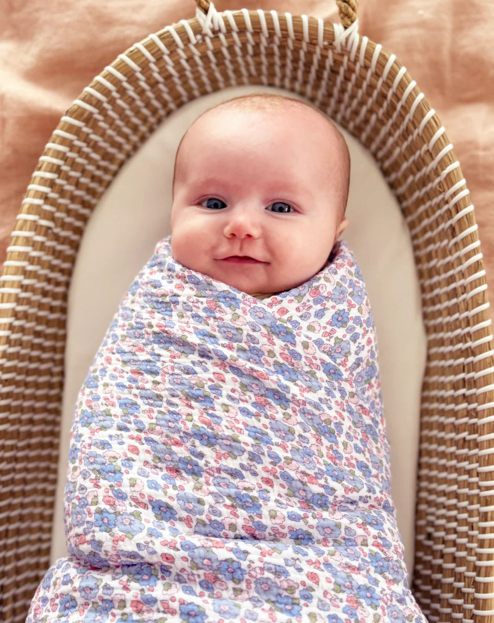 Alimrose Muslin Swaddle Liberty Blue. Baby swaddled in a floral blanket in a woven basket
