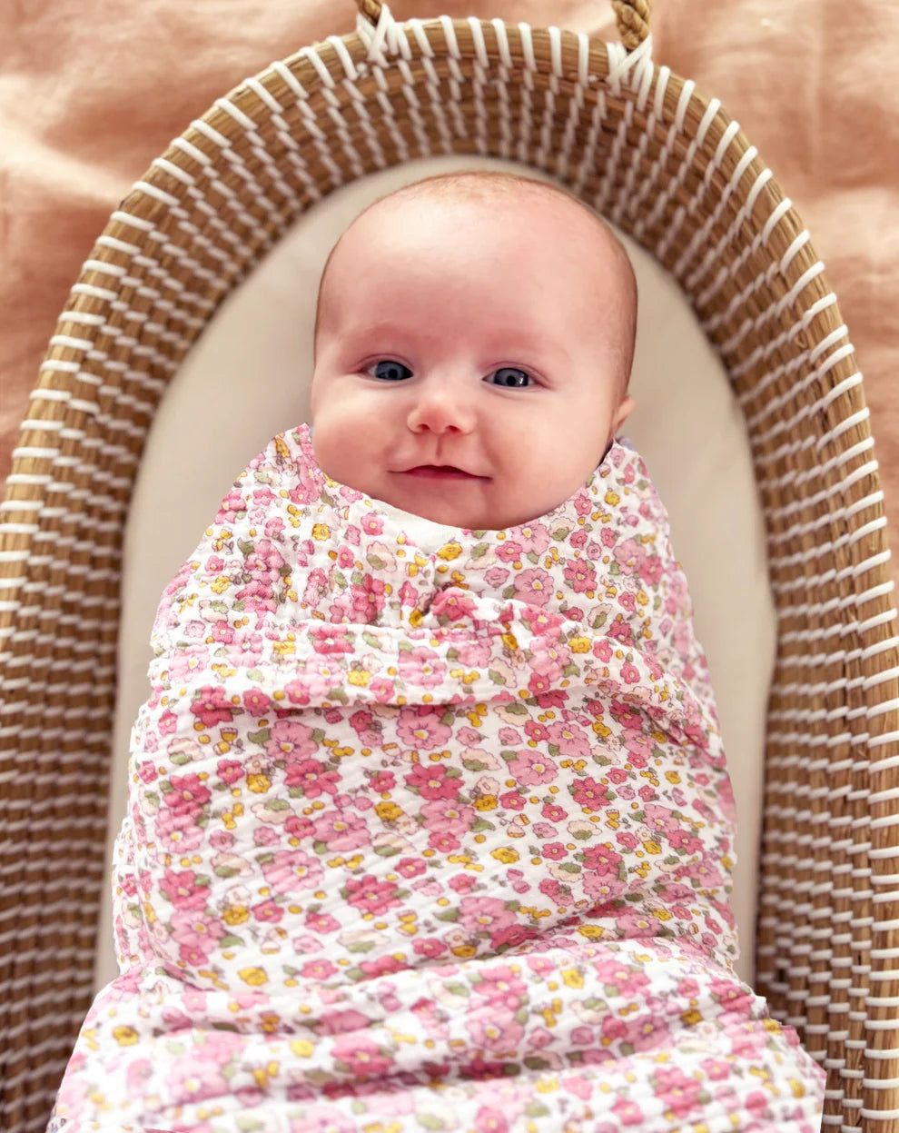 Alimrose Muslin Swaddle Rose Garden. Baby wrapped in a floral swaddle blanket sitting in a woven basket.