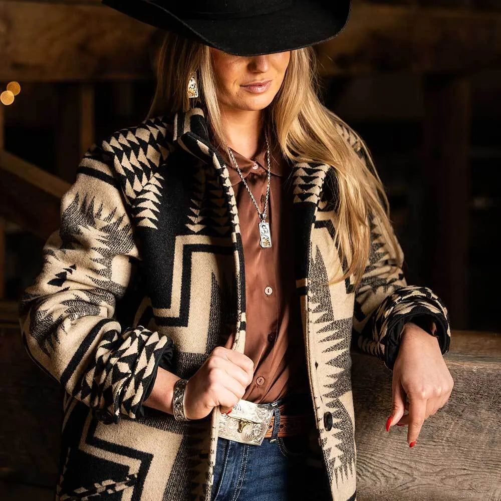 A woman wearing a black and beige geometric patterned jacket, a brown button-down shirt, blue jeans, and a black cowboy hat, posed in a rustic indoor setting.