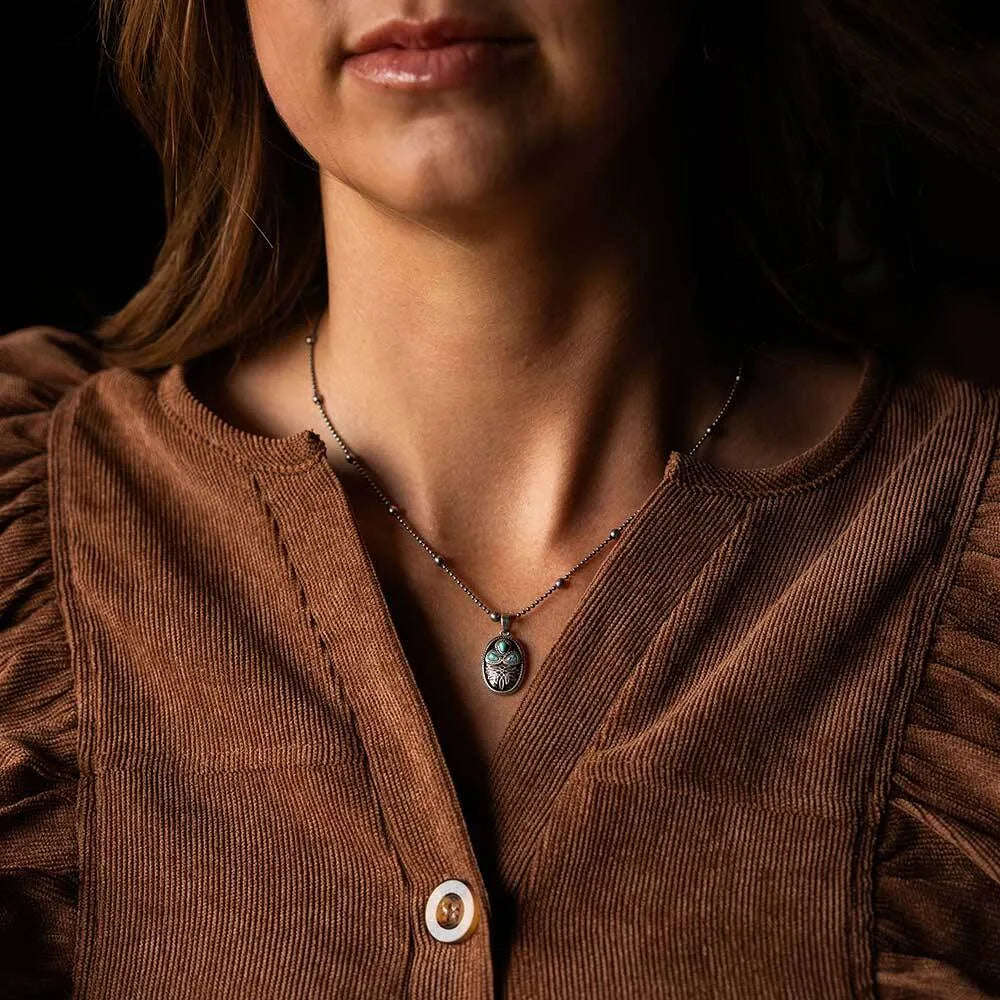 A close-up lifestyle shot of a woman wearing a silver-toned beaded necklace with a small oval pendant, paired with a brown corduroy top against a dark background.