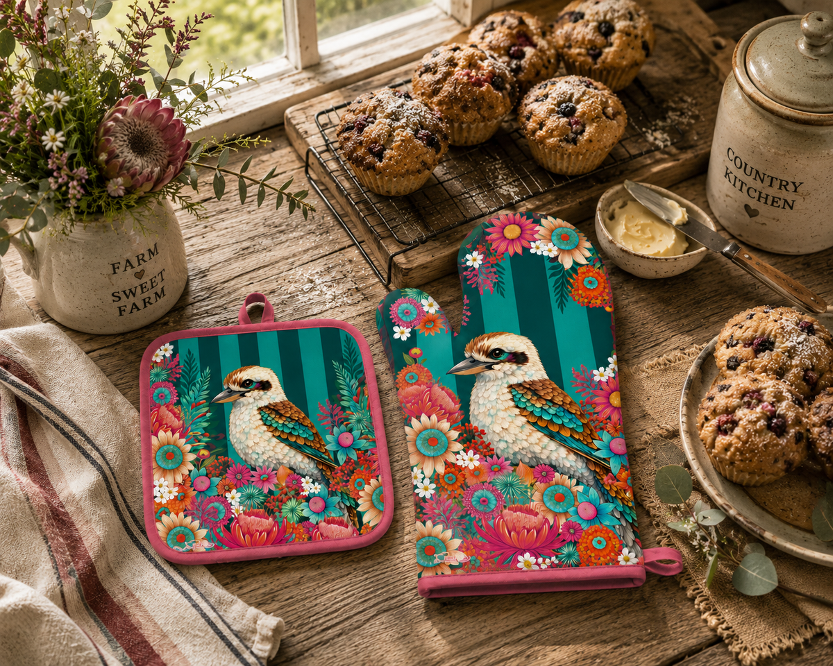 Two kitchen oven mitts with a colorful kookaburra and floral design are displayed on a rustic wooden table alongside muffins, a jar, and a striped towel. The mitts feature a bright pink border and a teal background with a detailed bird and floral pattern.