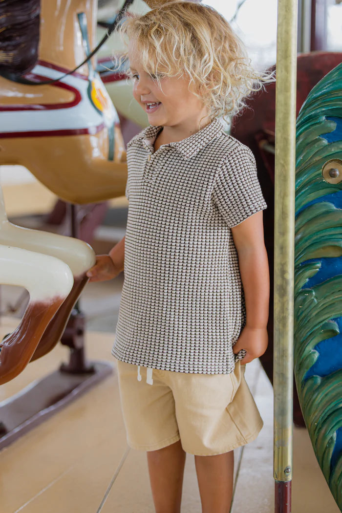 Milky Biscuit Short. Child in a checkered shirt and shorts standing near a carousel.