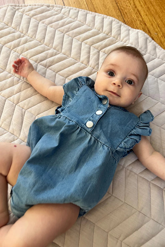 Milky Denim Frill Playsuit. Baby wearing a blue denim romper lying on a quilted surface