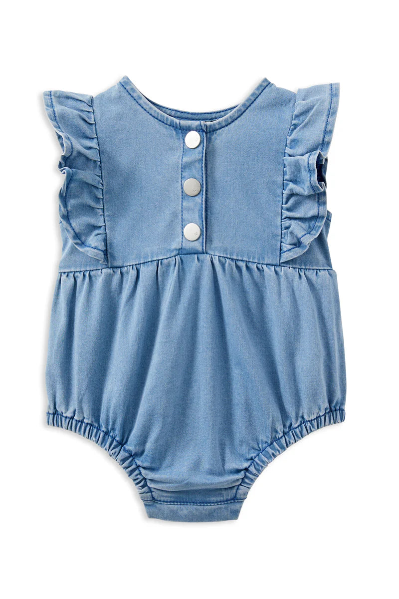 Milky Denim Frill Playsuit. Baby wearing a blue denim romper lying on a quilted surface