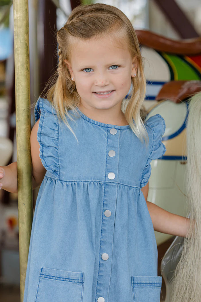 Milky Girl's Denim Frill Dress. Child wearing a blue denim dress holding a broom indoors.
