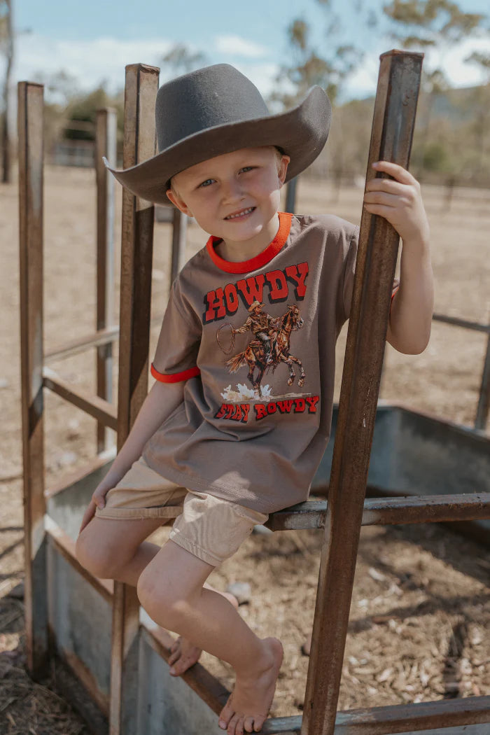 Milky Stay Rowdy Tee. Child wearing a cowboy hat and &#39;Howdy&#39; shirt sitting on a wooden structure outdoors.